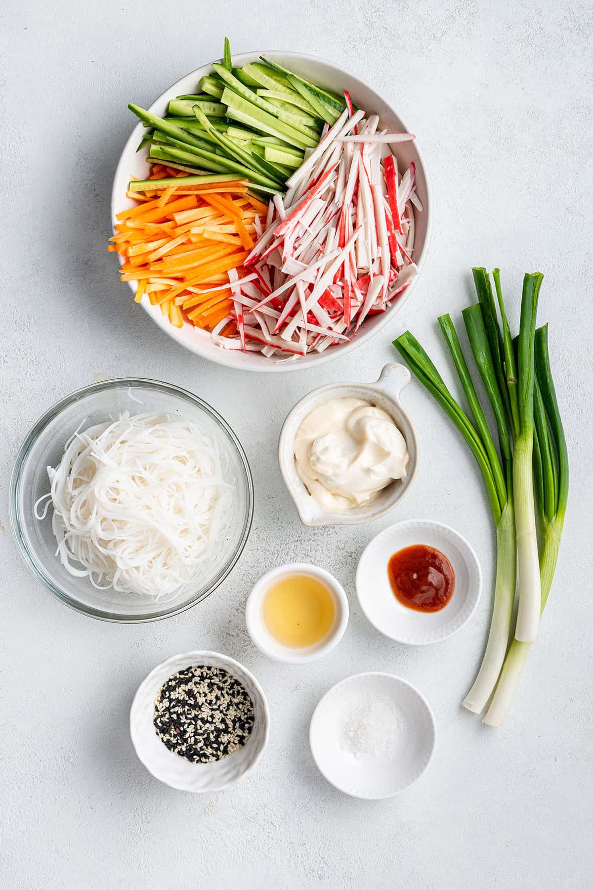 Overhead view of the ingredients needed for kani salad: a bowl of sliced carrots, cucumbers, and imitation crab meat, a bowl of cold rice noodles, a bowl of mayonnaise, a bowl of sriracha, a bowl of apple cider vinegar, a bowl of salt, a bowl of sesame seeds, and some scallions