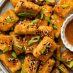 Overhead view of a plate of baked tofu, topped with scallions and sesame seeds, next to a bowl with a dipping sauce