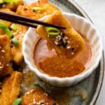 Close up of a pair of chopsticks dipping a piece of tofu, garnished with sesame seeds and scallions, into a dipping sauce, with more pieces in the background