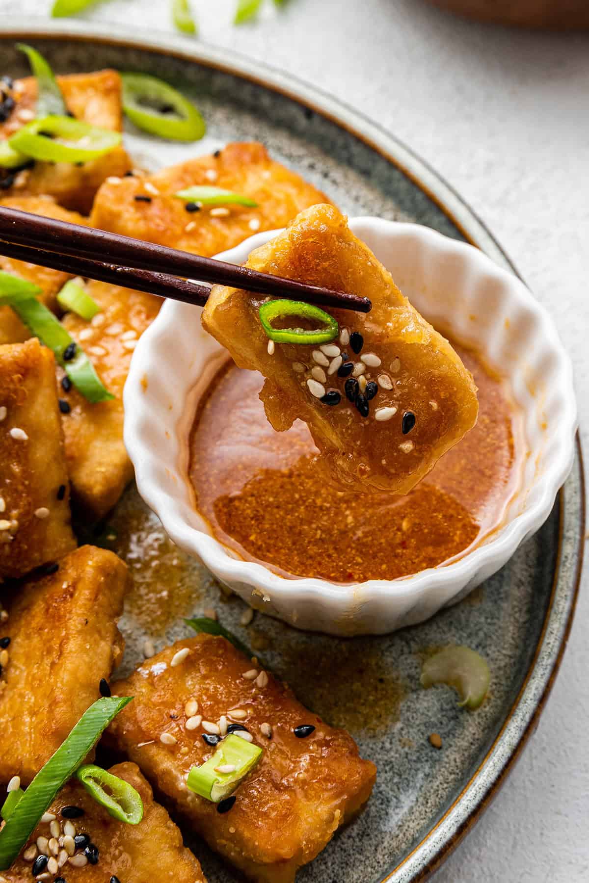 Close up of a pair of chopsticks dipping a piece of tofu, garnished with sesame seeds and scallions, into a dipping sauce, with more pieces in the background