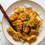 A plate with a pile of baked tofu on top of rice, garnished with scallions and sesame seeds, with chopsticks in it