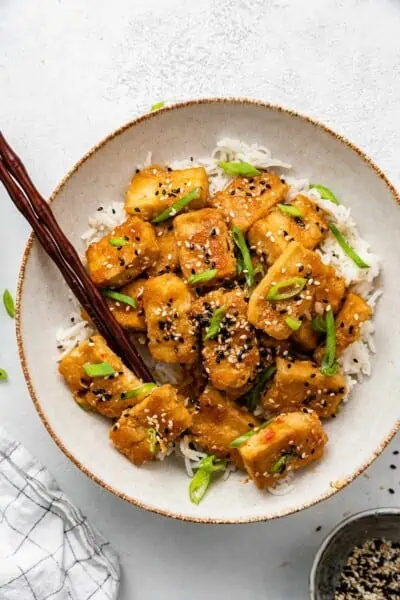 A plate with a pile of baked tofu on top of rice, garnished with scallions and sesame seeds, with chopsticks in it