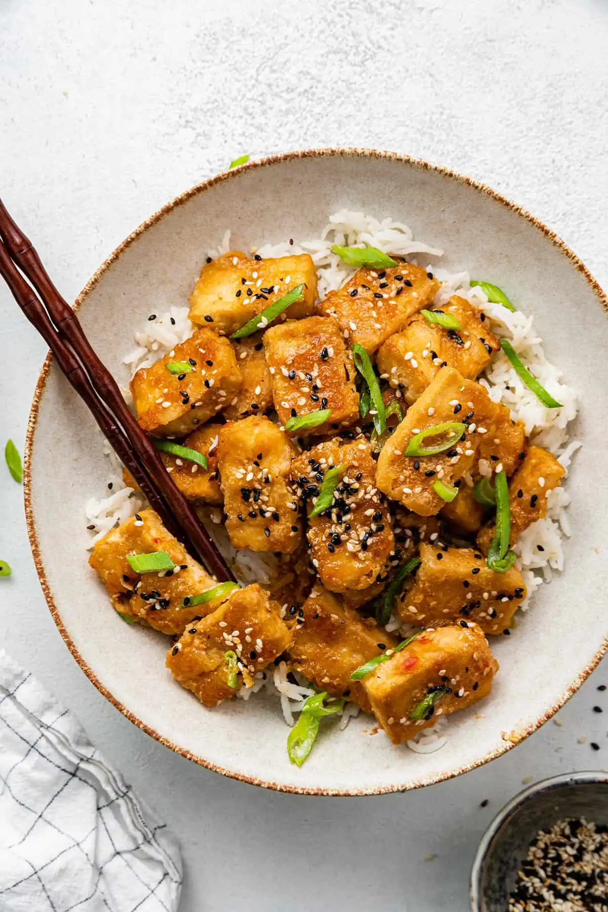 This Korean tofu recipe is crispy on the outside, juicy and tender on the inside, and marinated in a sweet and spicy sauce. A plate with a pile of baked tofu on top of rice, garnished with scallions and sesame seeds, with chopsticks in it