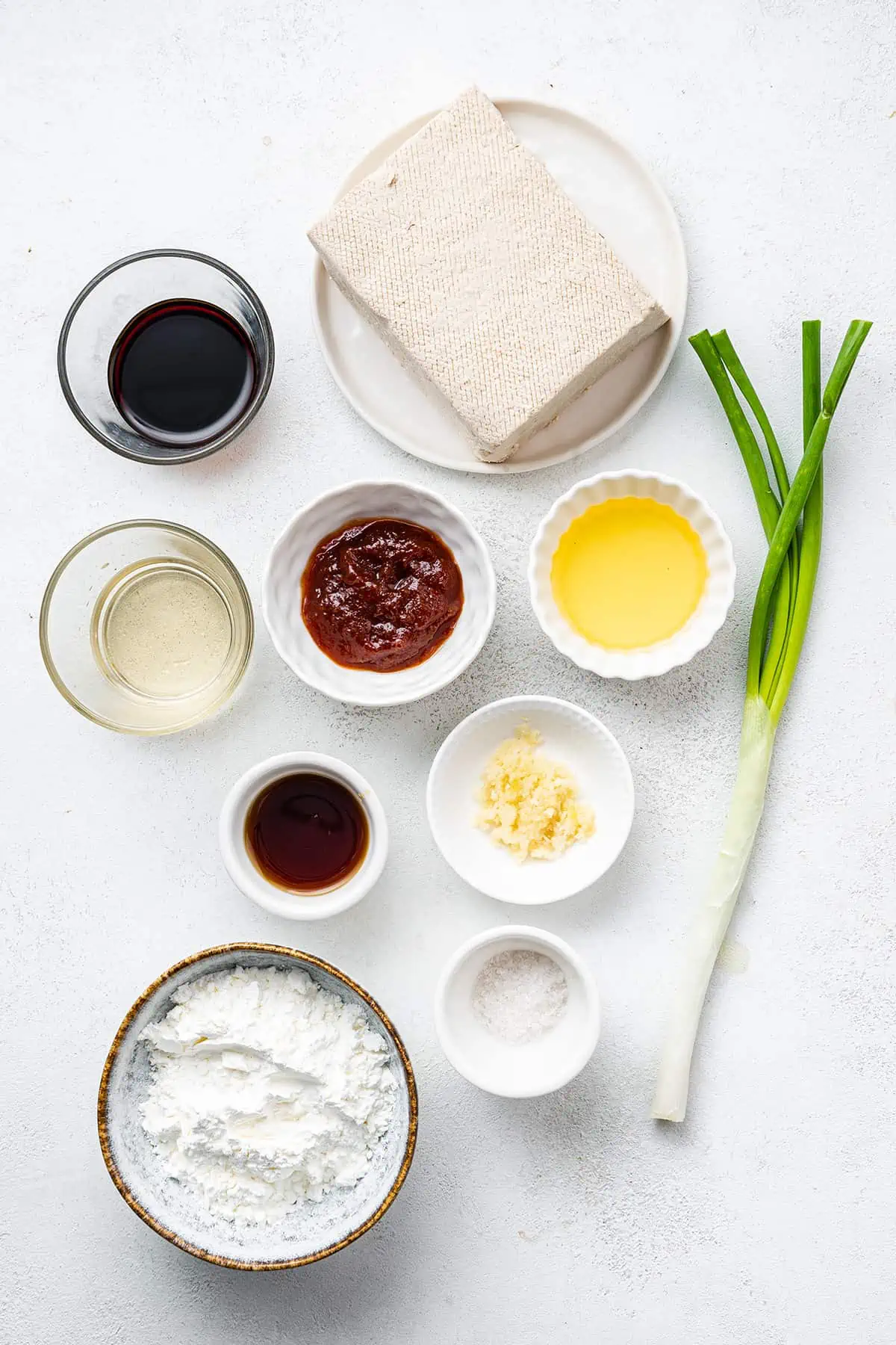 Overhead view of the ingredients needed for Korean tofu: a block of tofu, a green onion, a bowl of soy sauce, a bowl of vinegar, a bowl of gochujang, a bowl of sesame oil, a bowl of garlic, a bowl of salt, a bowl of cornstarch, and a bowl of maple syrup