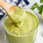 A jar of cilantro lime sauce with a spoon being removed from it, surrounded by lime slices, cilantro leaves, and a kitchen towel