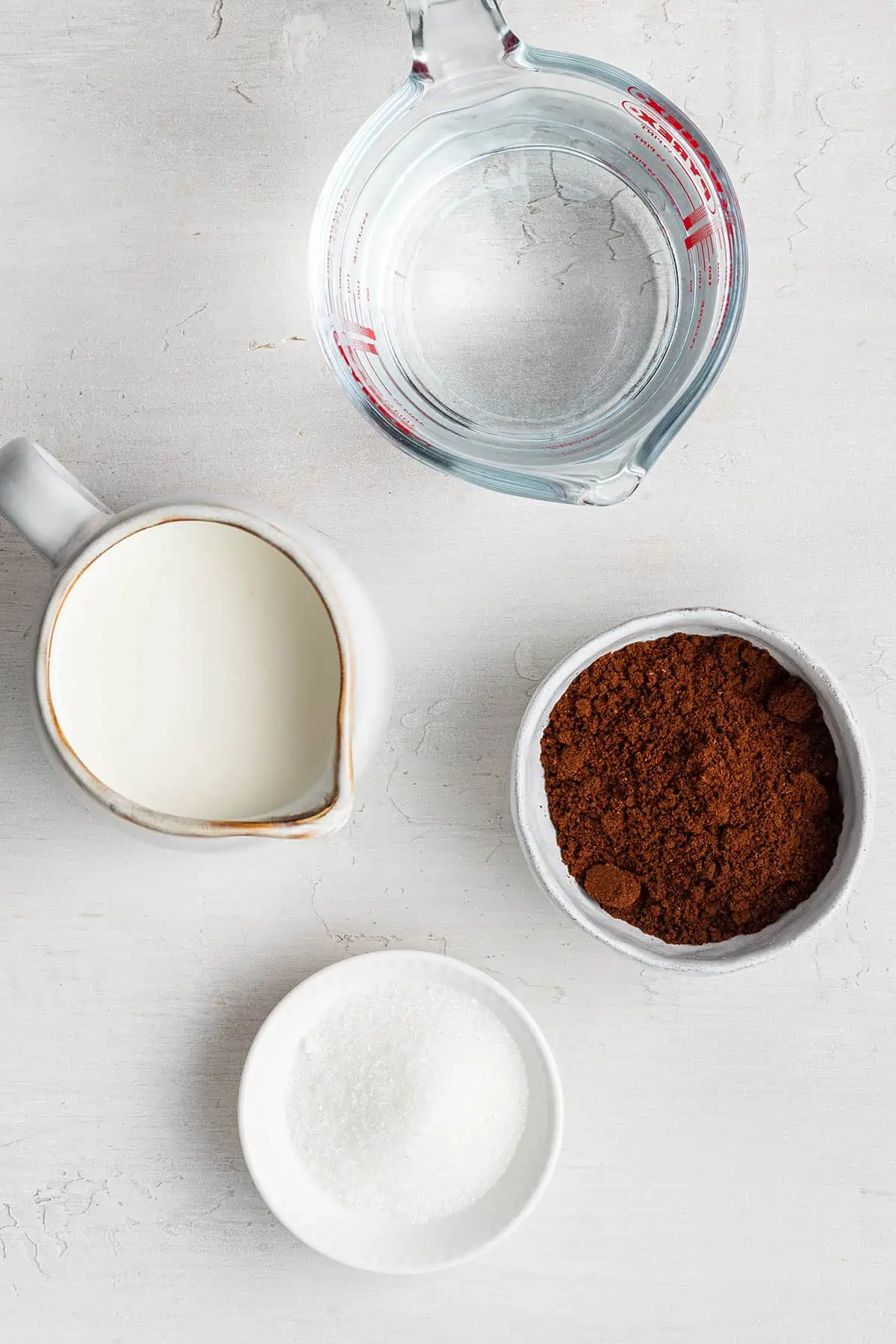 Overhead view of the ingredients needed for cortaditos: a bowl of coffee grounds, a bowl of sugar, a pitcher of evaporated milk, and a pyrex of water