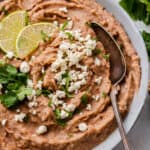 Close up of a bowl of refried bean stopped with cheese, cilantro, and lime slices, with a spoon in it