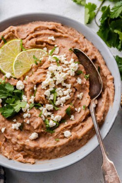 Close up of a bowl of refried bean stopped with cheese, cilantro, and lime slices, with a spoon in it