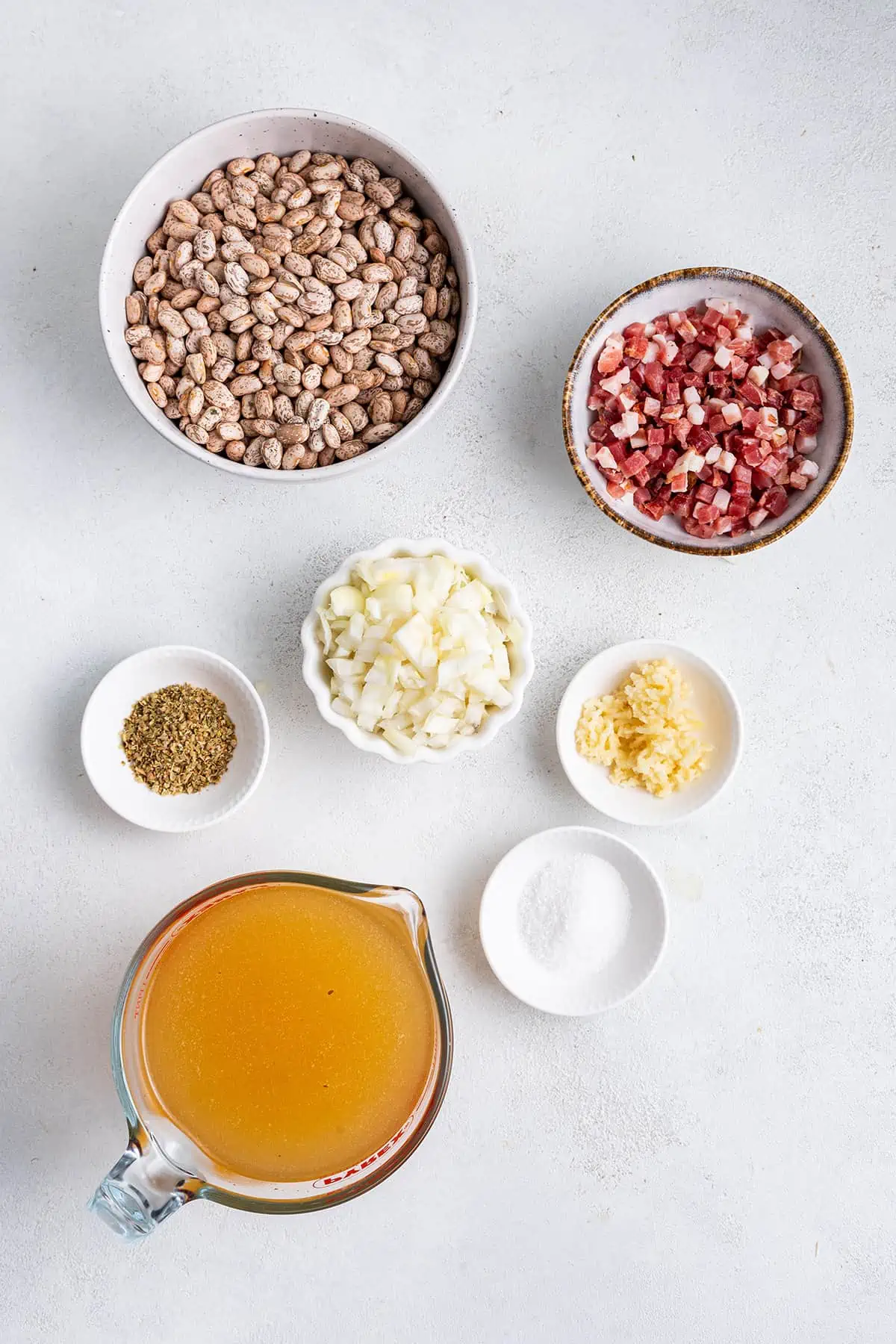 Overhead view of the ingredients needed for Instant Pot refried beans: A bowl of dried pinto beans, a bowl of diced raw bacon, a bowl of diced onions, a bowl of minced garlic, a bowl of salt, a bowl of oregano, and a pyrex of chicken broth