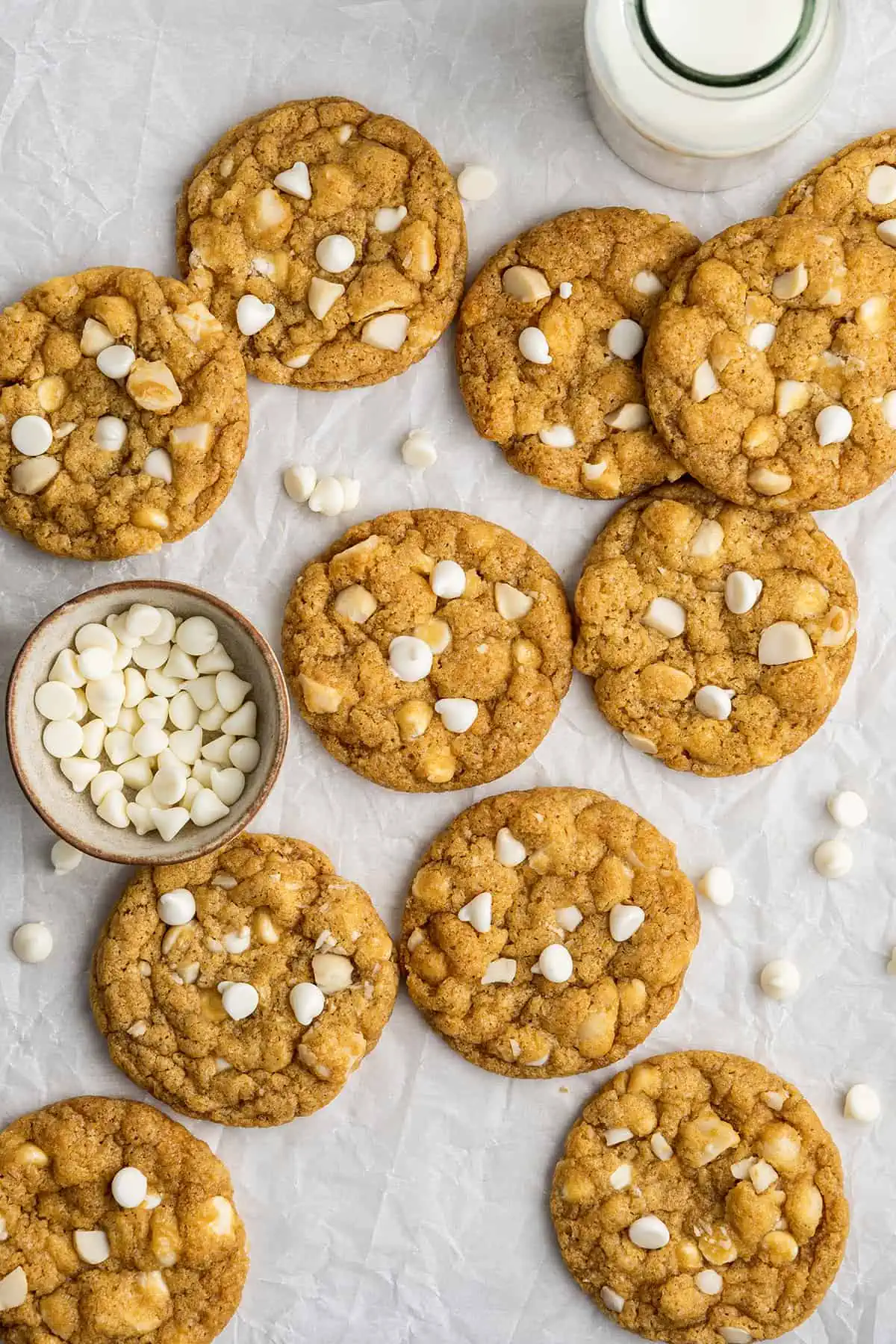 Overhead view of a bunch of white chocolate chip and macadamia nut cookies, with a jar of milk and a bowl of white chocolate chips