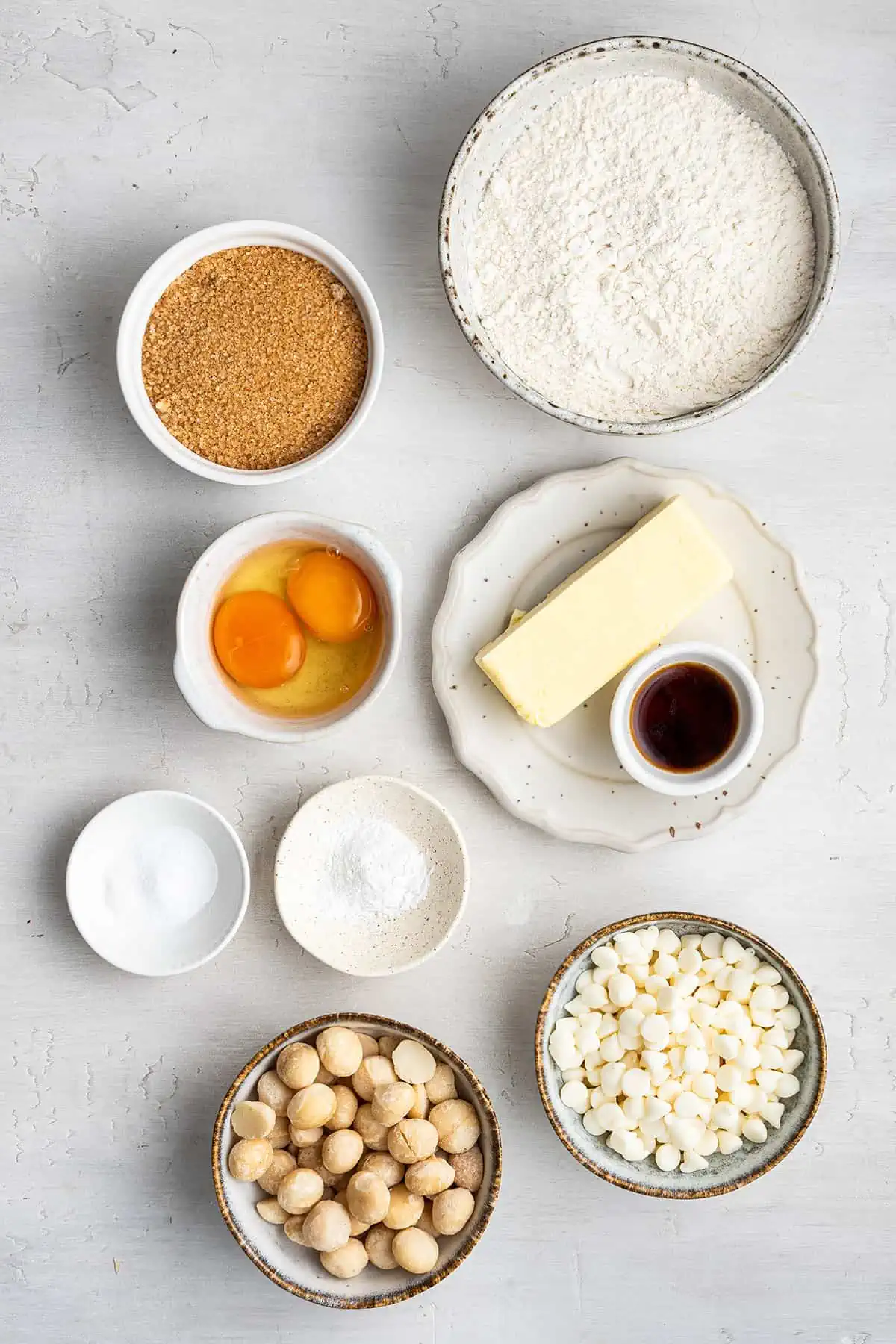 Overhead view of the ingredients needed for macadamia nut cookies: a bowl of flour, a bowl of raw sugar, a bowl of eggs, a bowl of baking powder, a bowl of baking soda, a bowl of white chocolate chips, a bowl of macadamia nuts, a bowl of vanilla extract, and a stick of butter