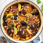 Overhead view of a dutch oven filled with chicken thighs, slivered almonds, lemon slices, green olives, dried apricots, and prunes, next to some silverware and a bowl of slivered almonds