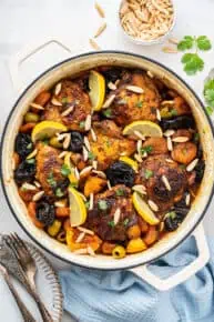 Overhead view of a dutch oven filled with chicken thighs, slivered almonds, lemon slices, green olives, dried apricots, and prunes, next to some silverware and a bowl of slivered almonds