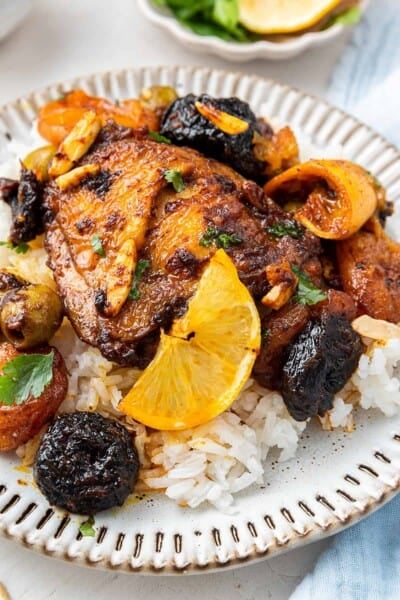 A plate with a chicken thigh on top of rice, topped with a lemon slice and cooked dried apricots and prunes