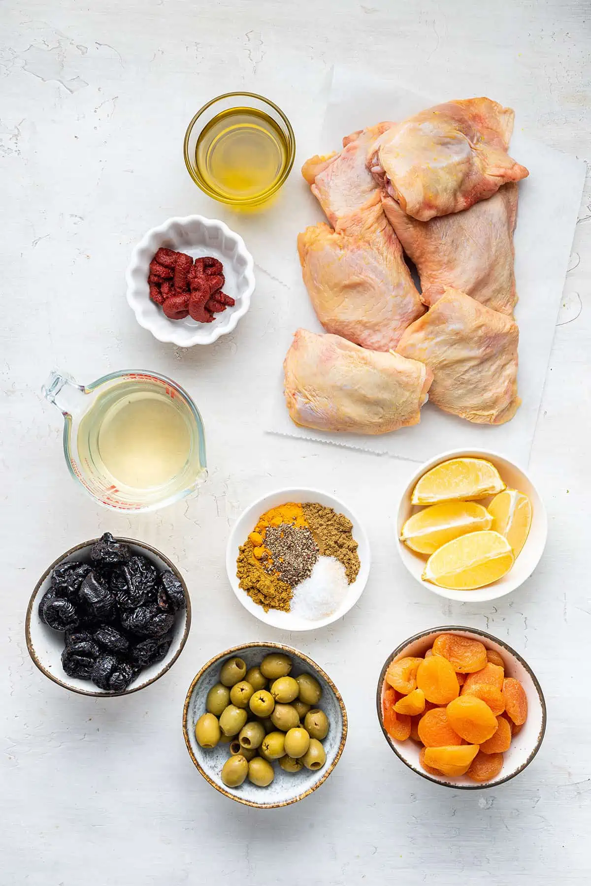 Overhead view of the ingredients needed for Moroccan chicken: skin-on chicken thighs, a bowl of olive oil, a bowl of tomato paste, a pyrex of white wine, a bowl of lemon slices, a bowl of spices, a bowl of dried apricots, a bowl of prunes, and a bowl of green olives
