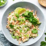 Overhead view of a slaw topped with cilantro and limes, next to a bowl of scallions, a lime slice, and a wooden spoon