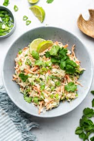 Overhead view of a slaw topped with cilantro and limes, next to a bowl of scallions, a lime slice, and a wooden spoon