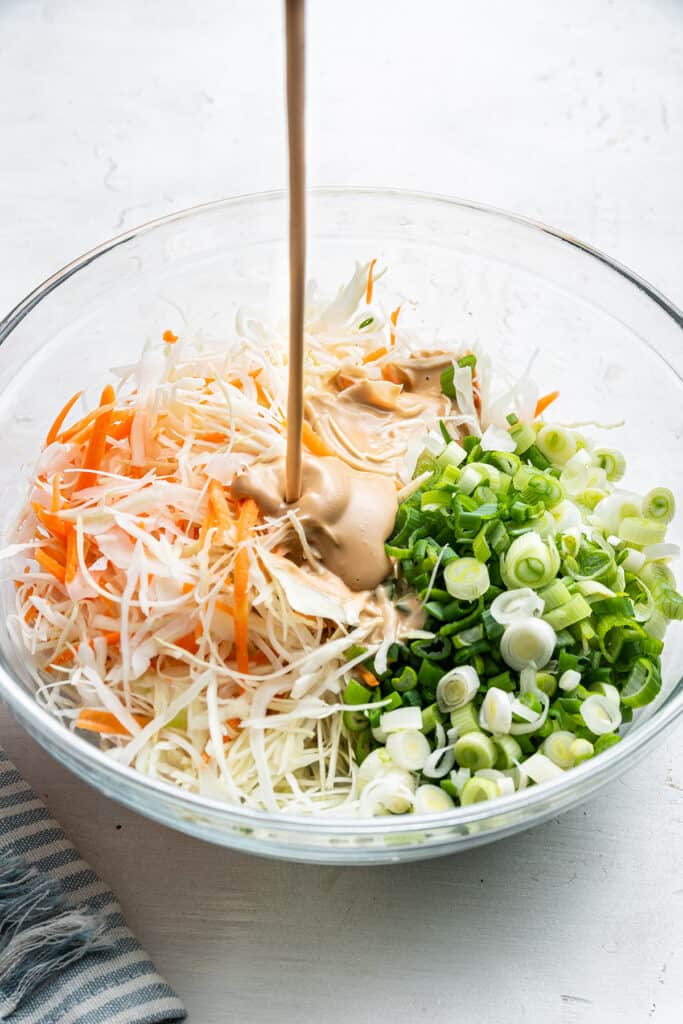 A bowl of cut up cabbage, carrots, and scallions, with a dressing being poured into it.