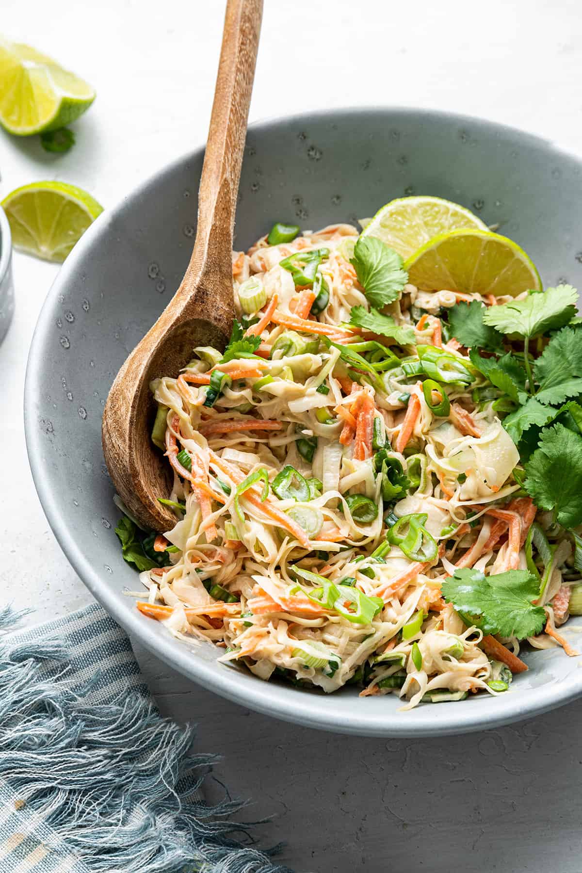 A bowl of slaw topped with cilantro and limes, with a wooden spoon in it.