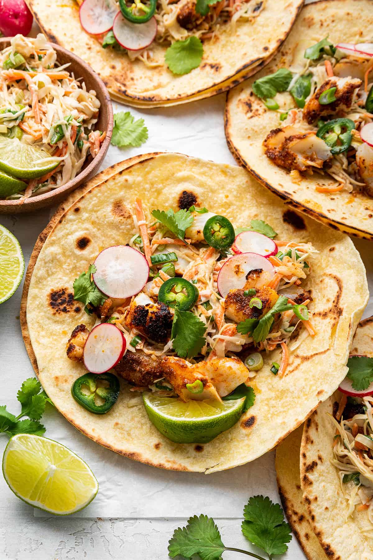 Overhead view of a lot of fish tacos next to each other, topped with lime, jalapeño, and radishes