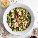 Overhead view of a bowl of broccoli cauliflower salad, next to a jar of dressing, a bowl of dried cranberries, and a bowl of sunflower seeds