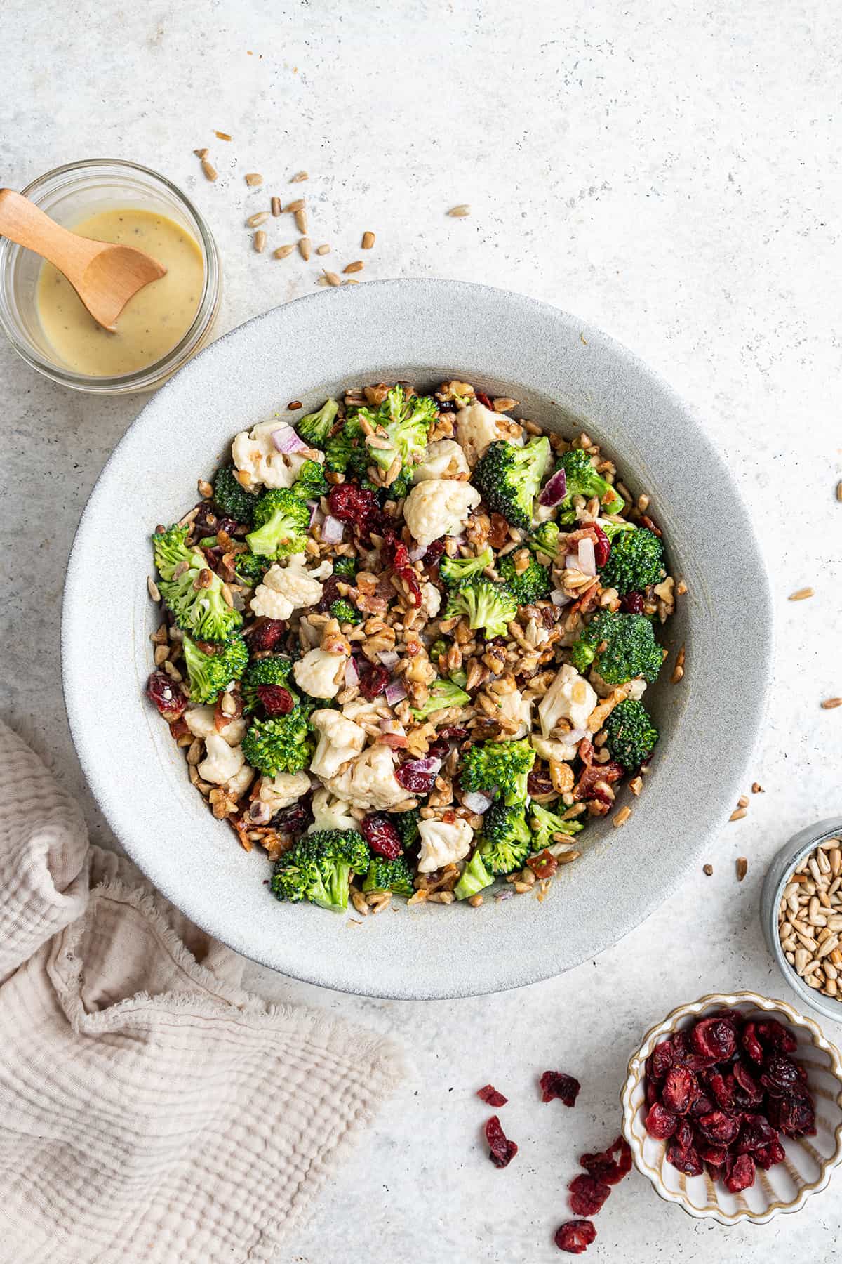 Overhead view of a bowl of broccoli cauliflower salad, next to a jar of dressing, a bowl of dried cranberries, and a bowl of sunflower seeds