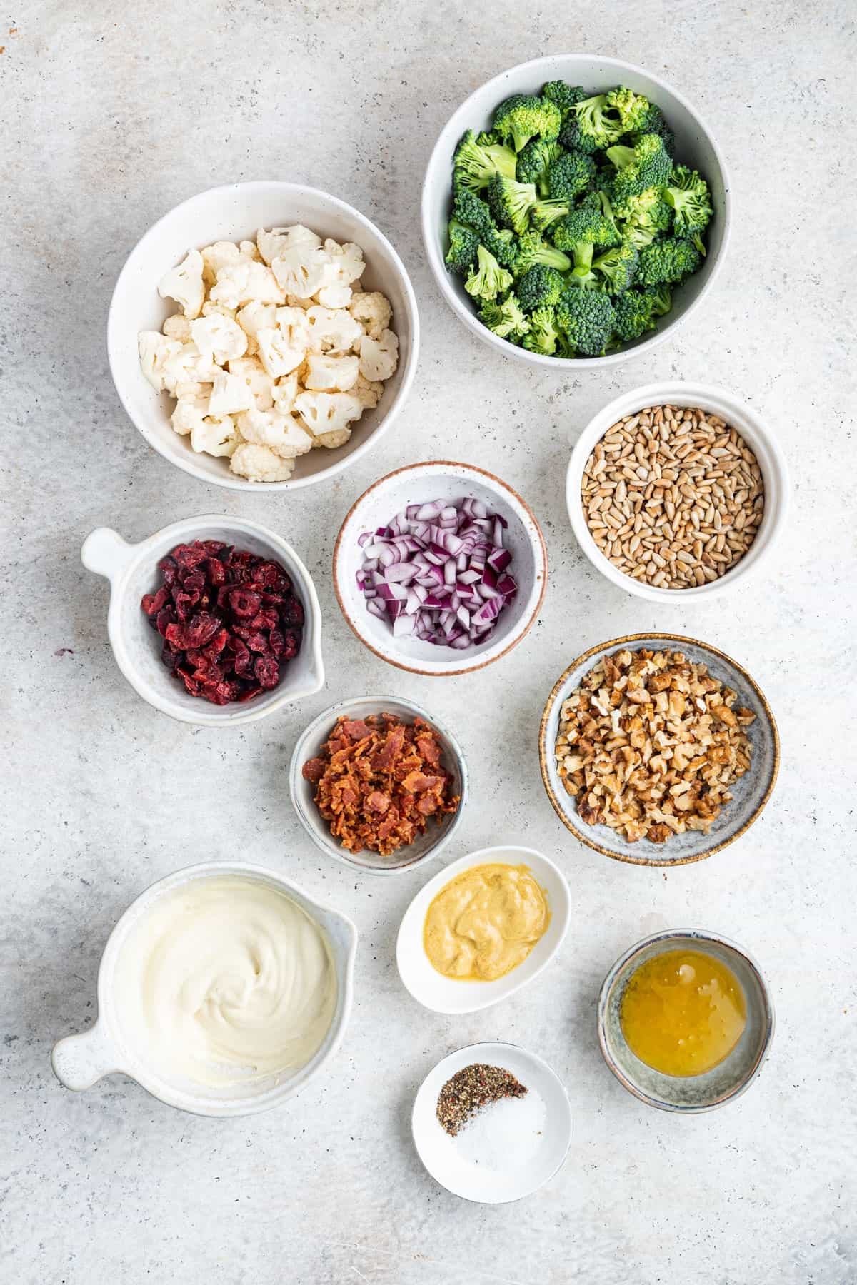 Overhead view of the ingredients needed for broccoli cauliflower salad: a bowl of broccoli, a bowl of cauliflower, a bowl of onions, a bowl of sunflower seeds, a bowl of walnuts, a bowl of dried cranberries, a bowl of bacon bits, a bowl of mayonnaise, a bowl of mustard, a bowl of honey, and a bowl of salt and pepper
