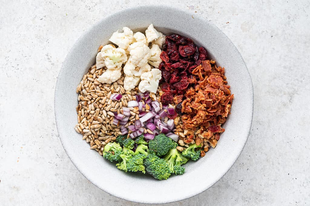 Overhead view of a bowl with broccoli, cauliflower, red onion, dried cranberries, sunflower seeds, walnuts, and bacon