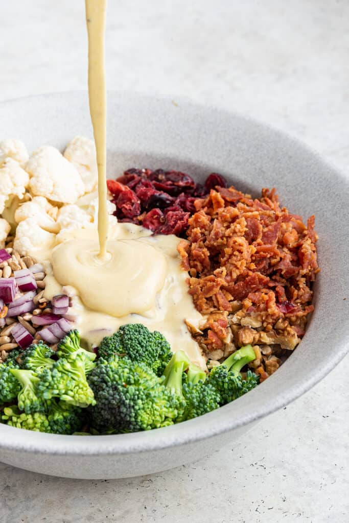 Mayo-based dressing being poured into a bowl with broccoli, onions, cauliflower, bacon, and dried cranberries