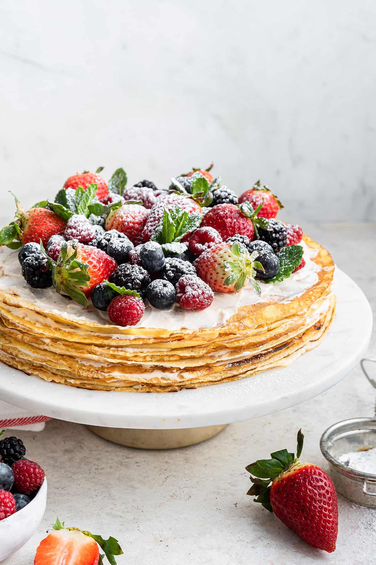 A crepe cake on a cake stand, topped with berries, powdered sugar, and mint