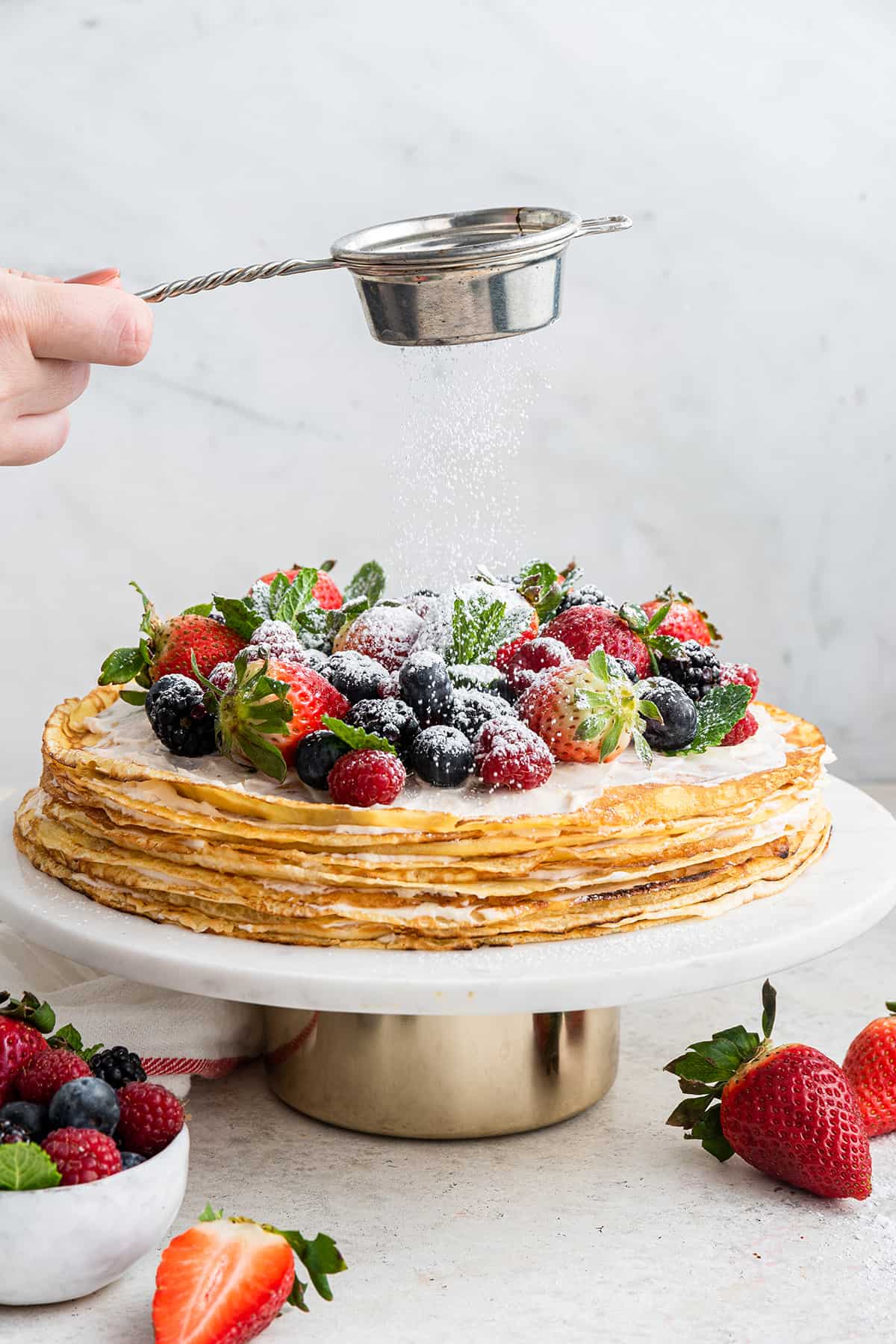 A hand dusting a crepe cake topped with berries with powdered sugar