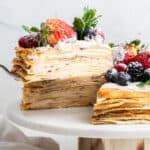 A spatula taking a slice of crepe cake topped with berries and mint off of a cake stand
