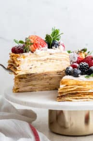 A spatula taking a slice of crepe cake topped with berries and mint off of a cake stand