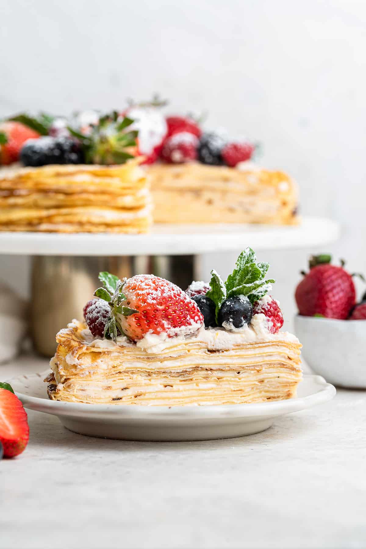 A plate with a slice of crepe cake covered in berries and mint, with whole crepe cake ion a cake stand in the background
