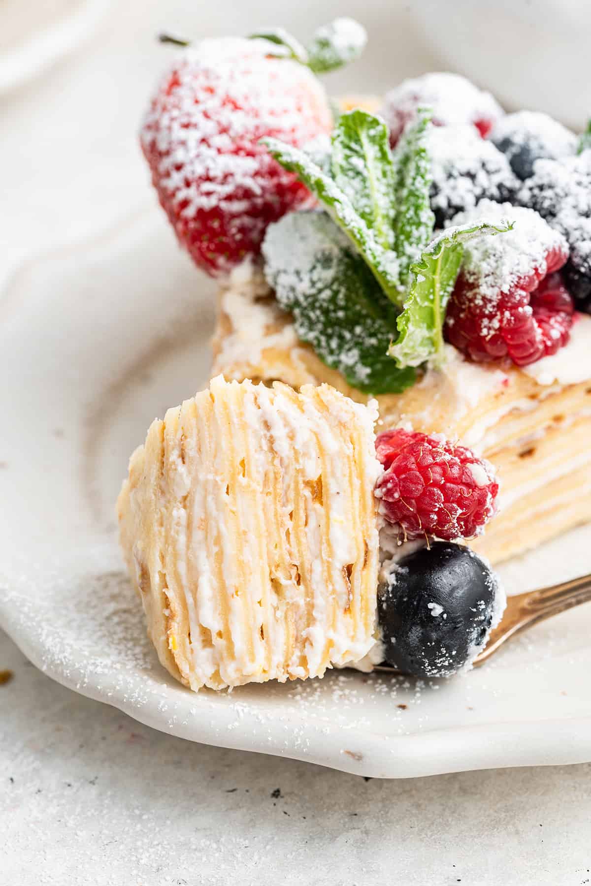 Close up of a slice of crepe cake topped with berries and mint, with a bite cut out