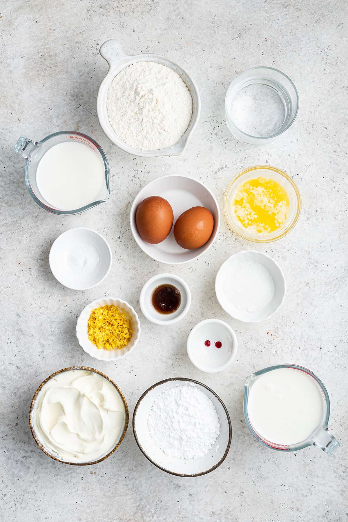 Overhead view of the ingredients needed for crepe cake: a bowl of flour, a glass of water, a bowl of milk, a bowl of eggs, a bowl of melted butter, a bowl of vanilla, a bowl of lemon zest, a bowl of sugar, a bowl of powdered sugar, a bowl of salt, a bowl of heavy cream, a bowl of mascarpone, and a bowl of red food dye.