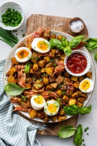 Overhead view of a plate of breakfast potatoes topped with bacon, basil, and soft boiled eggs, with a bowl of ketchup, a bowl of salt, and a bowl of chives