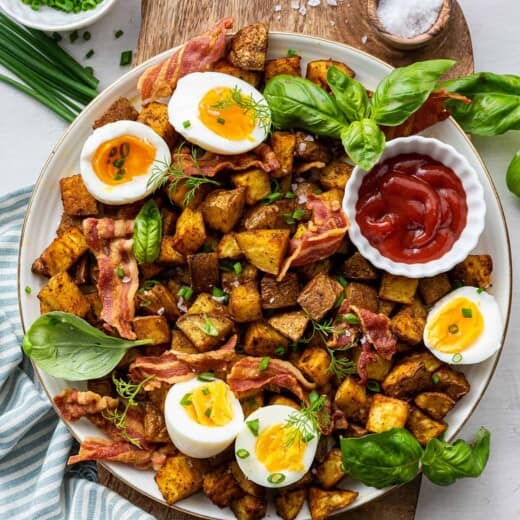 Overhead view of a plate of breakfast potatoes topped with bacon, basil, and soft boiled eggs, with a bowl of ketchup, a bowl of salt, and a bowl of chives