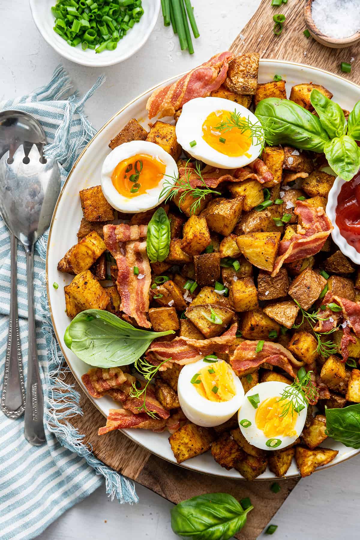 Overhead view of a plate of breakfast potatoes topped with bacon, basil, and soft boiled eggs