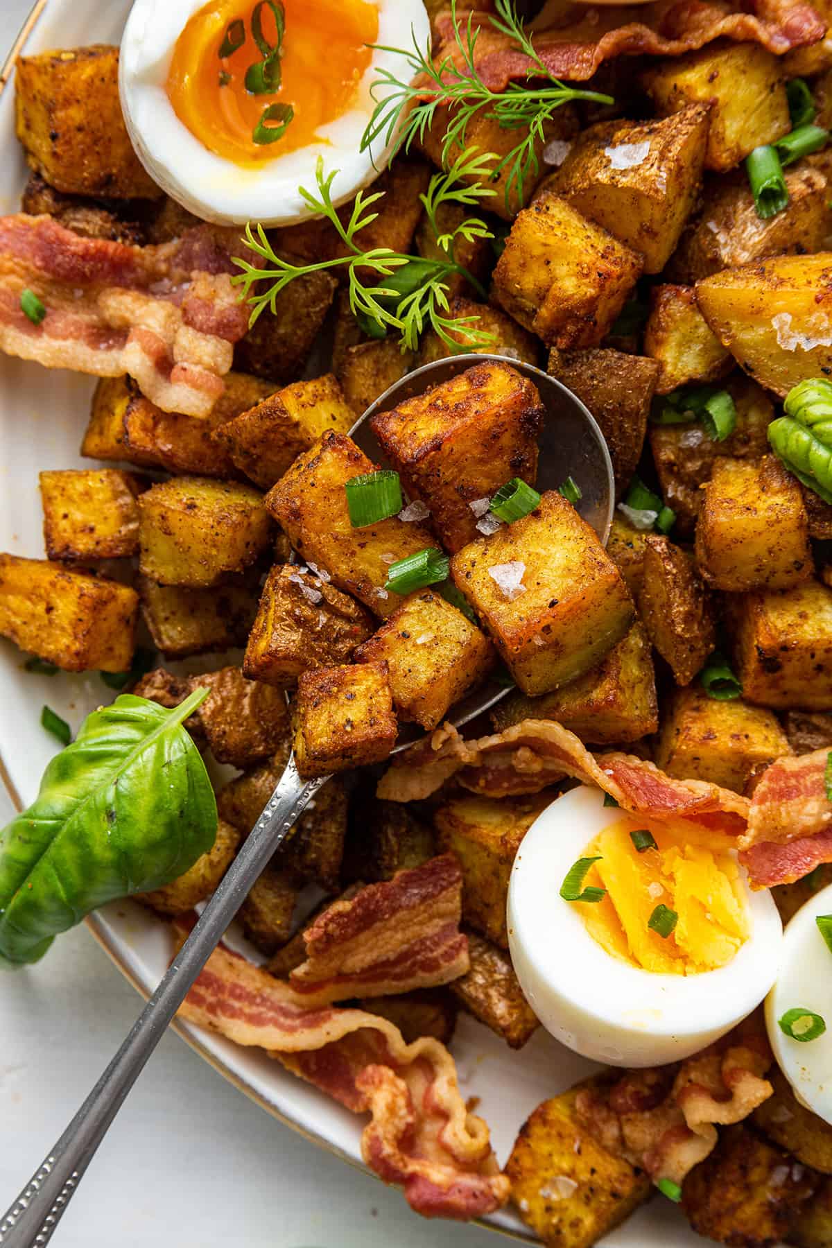 A spoon with a spoonful of roasted potatoes, on top of a plate full of potatoes, soft boiled eggs, and bacon.