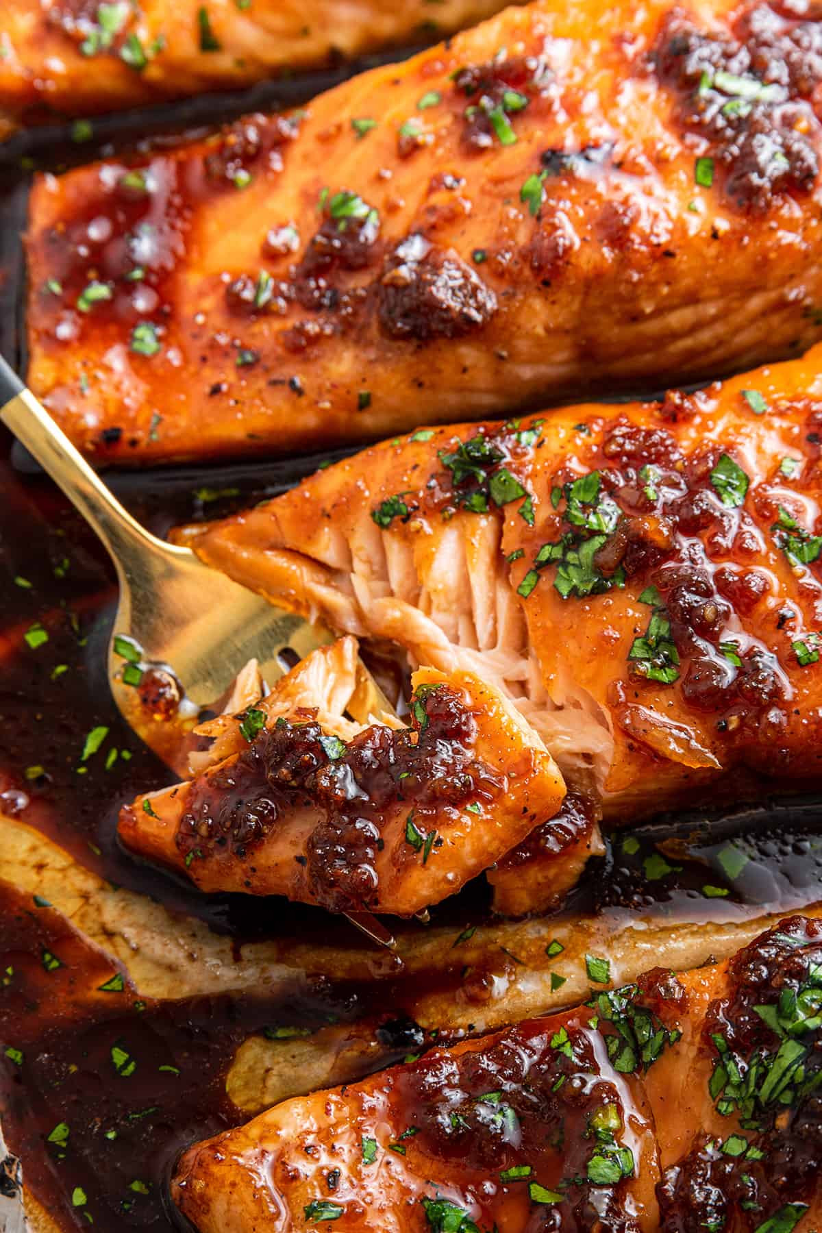Close up of a fork taking a bite out of a honey glazed salmon filet on a baking sheet, surrounded by more salmon fillets