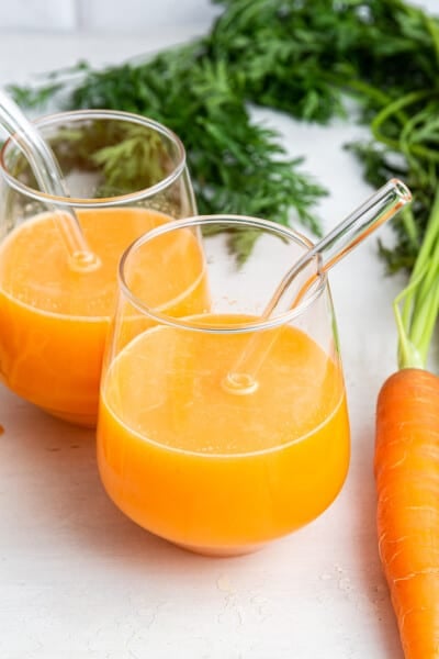 Two glasses of carrot juice with glass straws set next to carrot on counter