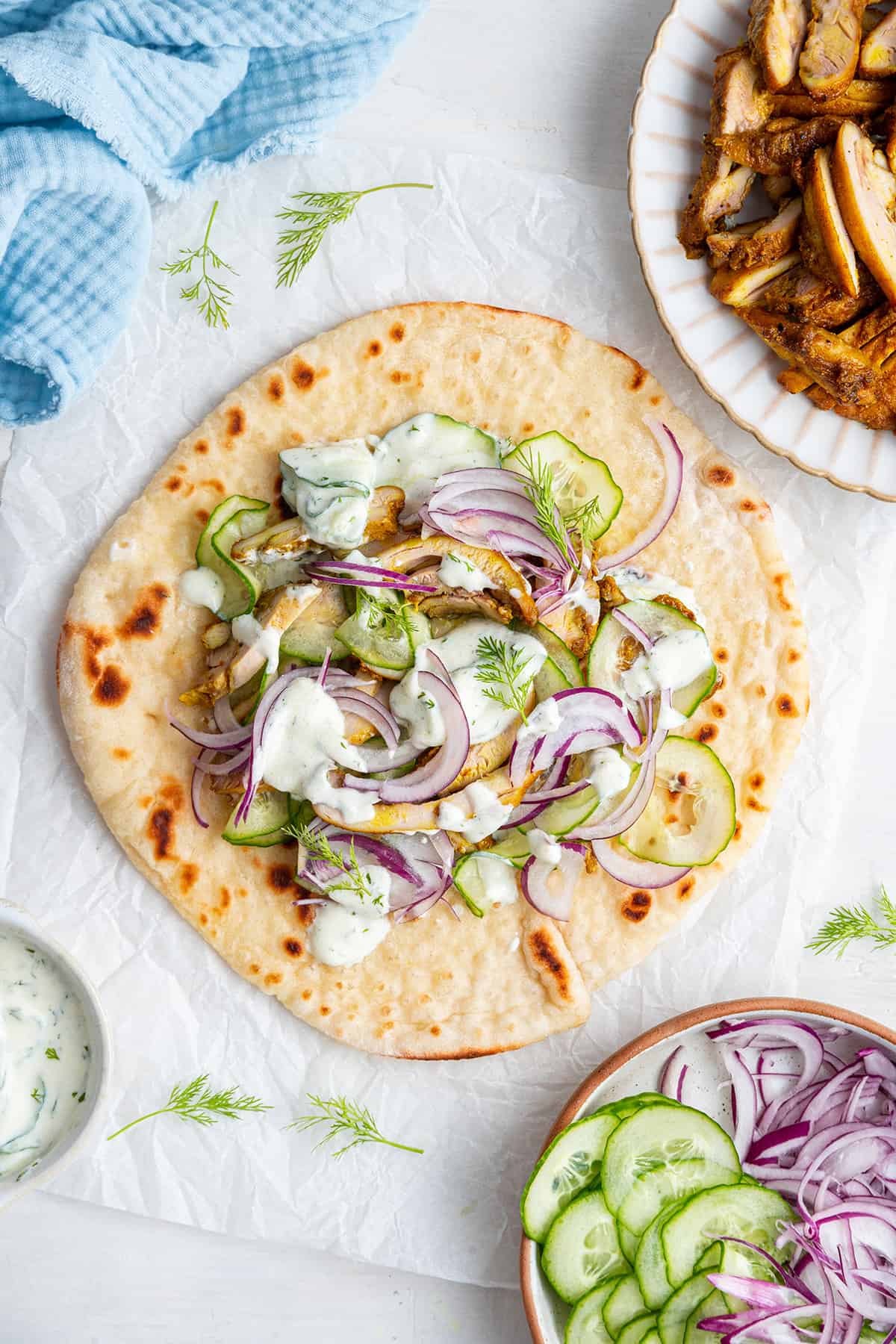 Overhead view of chicken shawarma gyro on parchment paper, with plate of chicken and toppings