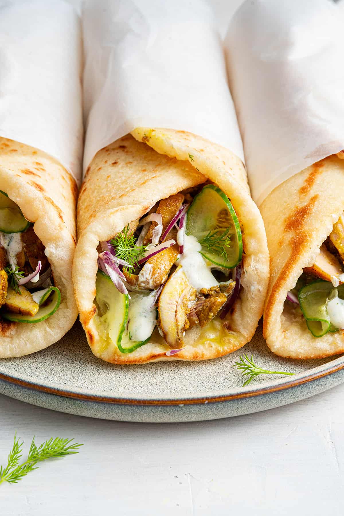 Three chicken shawarma gyros lined up on plate with scattered fronds of dill