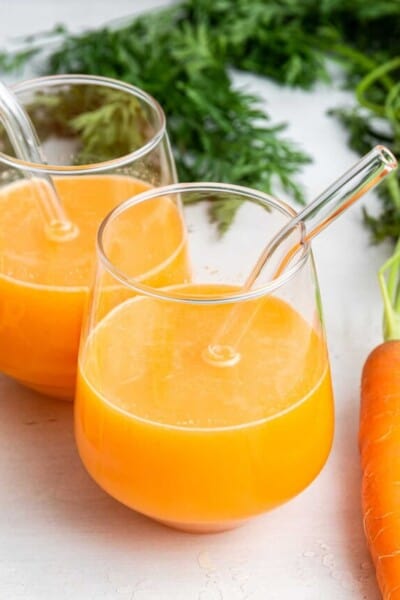 Two glasses of carrot juice with glass straws set next to carrot on counter