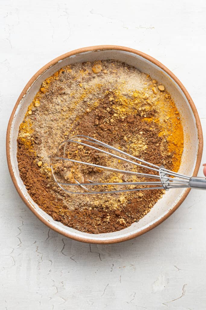 Overhead view of a whisk mixing together the ingredients for ras el hanout in a bowl