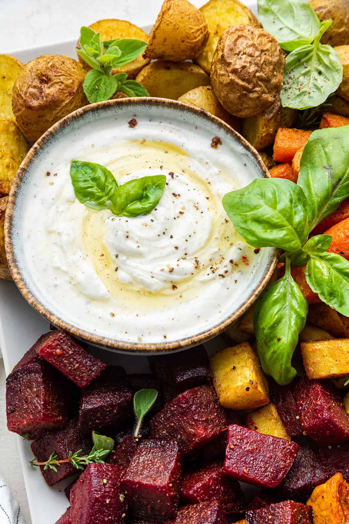 Bowl of Greek yogurt dip garnished with olive oil, pepper, and basil leaves, on platter of roasted veggies
