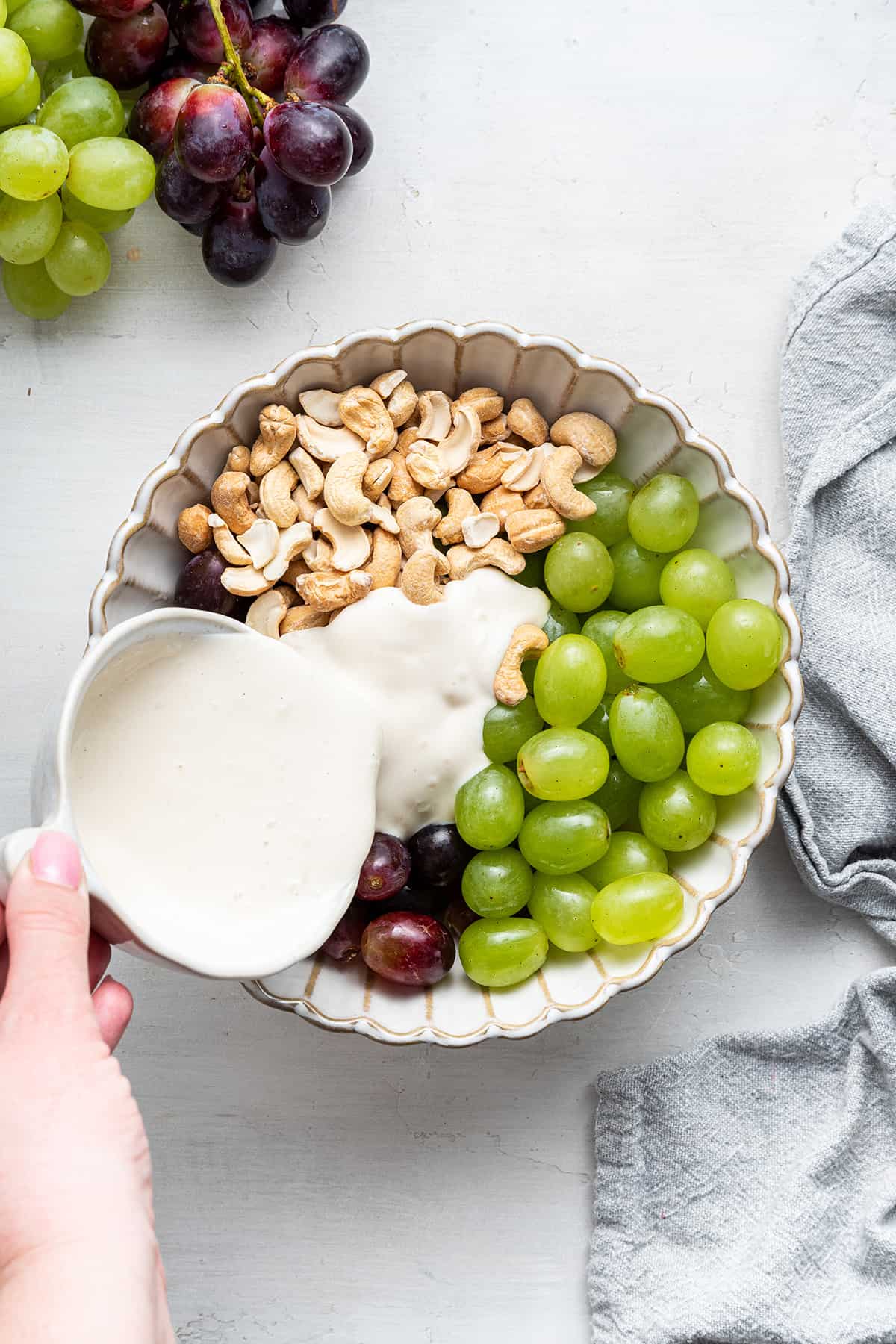 A pitcher pouring a yogurt dressing into a bowl with yellow grapes, red grapes, and cashews