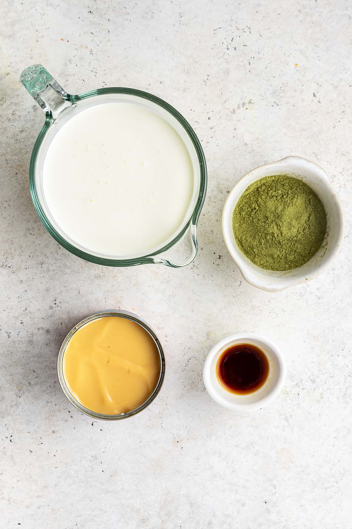 Overhead view of the ingredients needed for matcha ice cream: a bowl of vanilla extract, a bowl of matcha powder, a pyrex of whipping cream, and a can of condensed milk