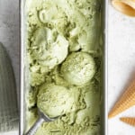 Overhead view of a loaf pan filled with matcha ice cream, with an ice cream scoop scooping a ball of ice cream